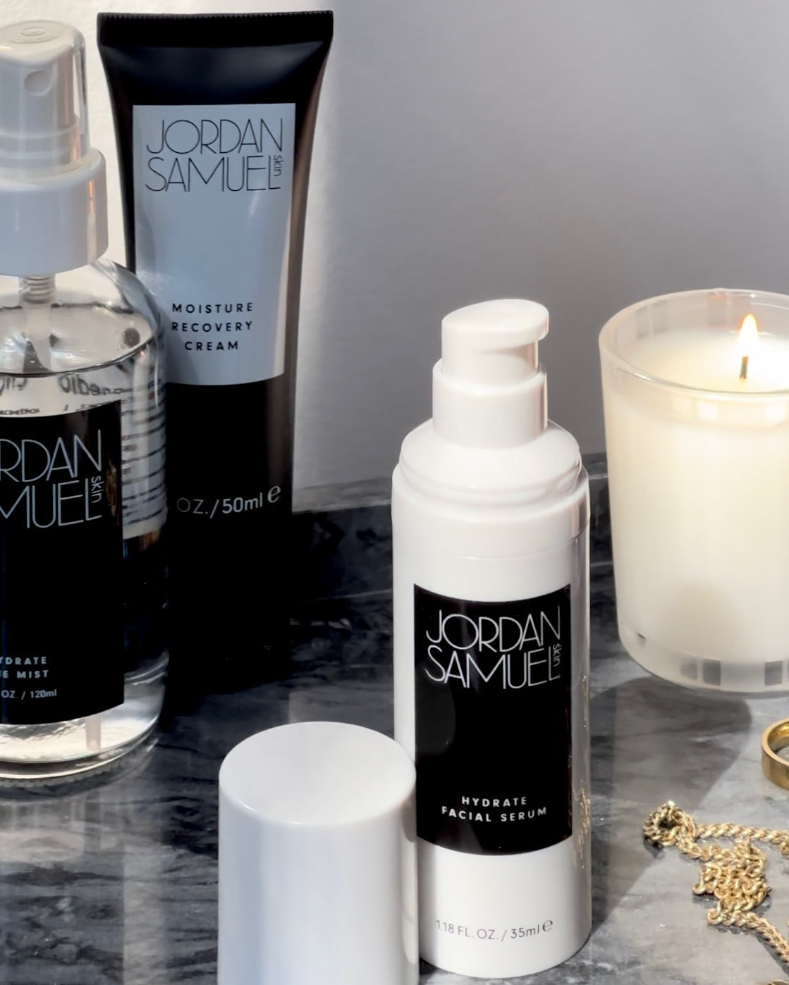 Hydrate Facial Serum's white pump bottle with black label stands in the foreground with gold jewelry. A lit white candle in a glass jar, the Moisture Recovery Cream's black tube with white label, and Hydrate the Mist's clear glass bottle with black label and white sprayer are seen in the background.