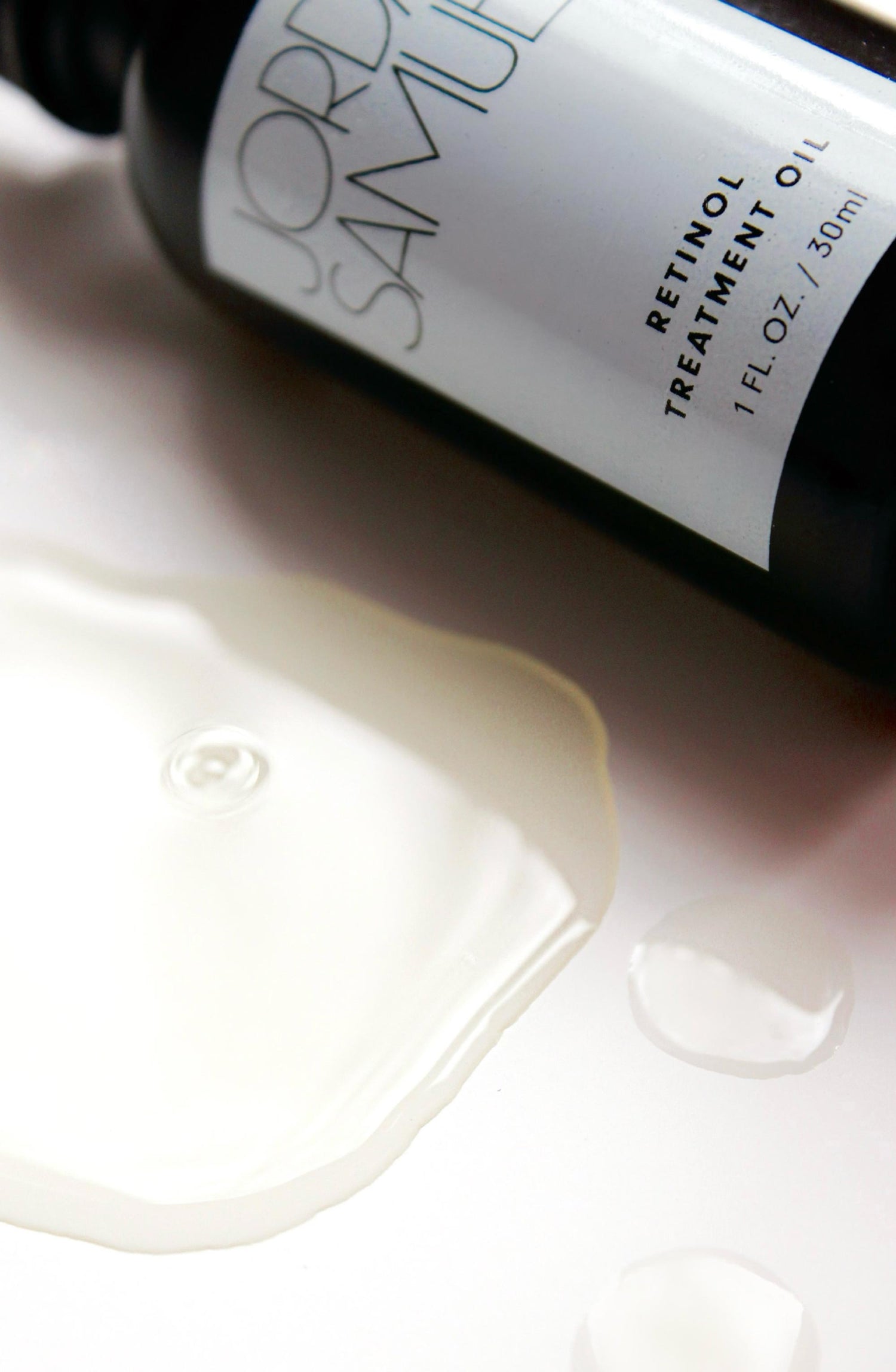 Closely cropped image of The Retinol Treatment Oil's black glass bottle with white label next to one large drop and two smaller droplets of the oil.