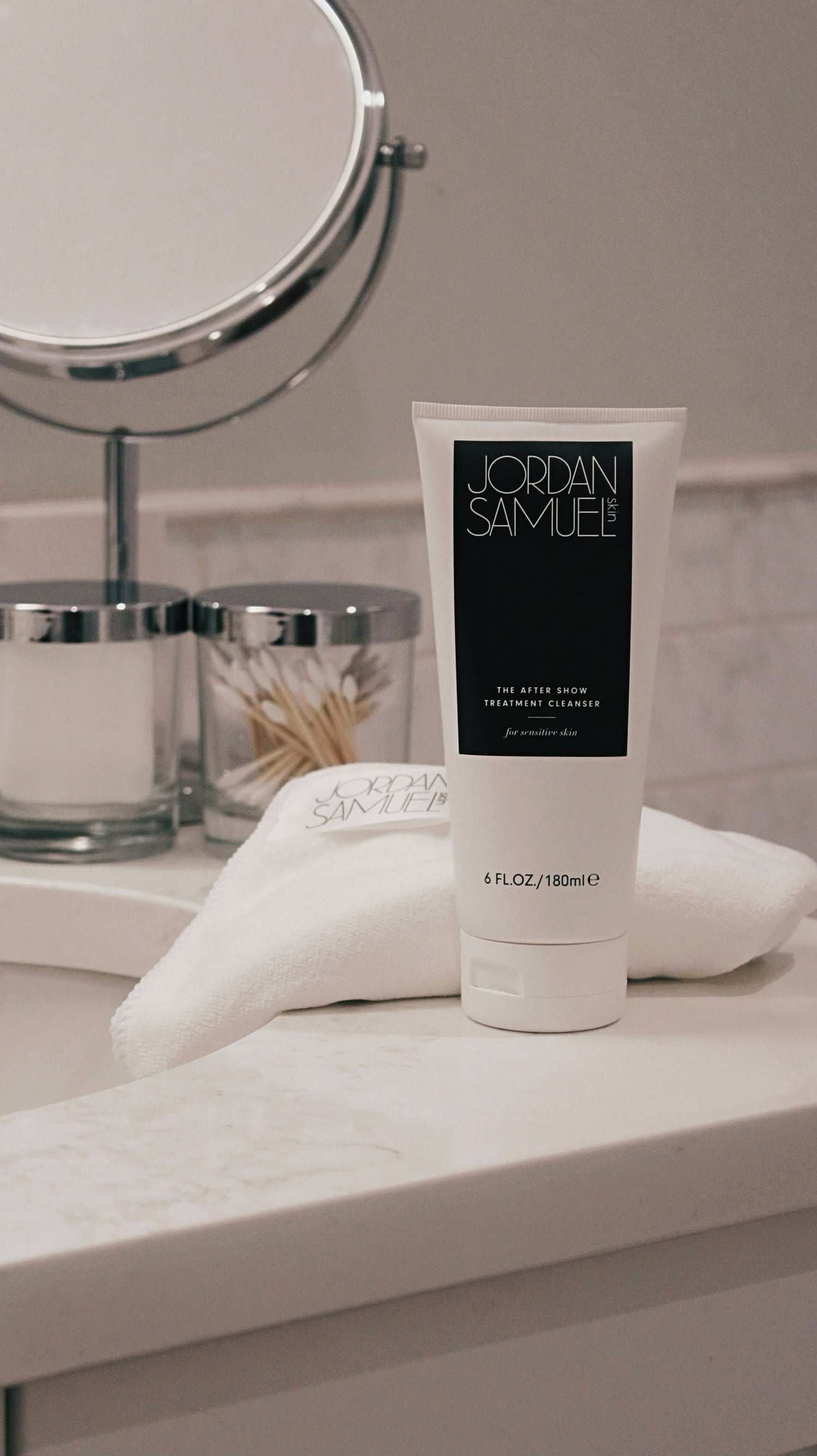 The After Show Treatment Cleanser for Sensitive Skin's white tube with black label stands on a white bathroom vanity in front of a branded white microfiber facial cloth, a round mirror and glass containers holding q-tips and cotton pads.