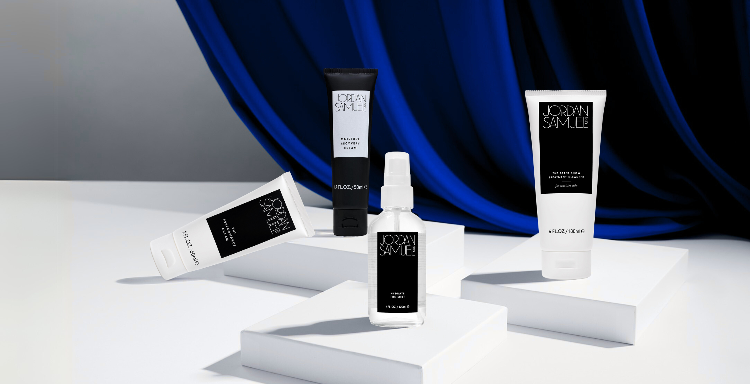 Image of The After Show Treatment Cleaner for Sensitive Skin's white tube with black label, Hydrate the Mist's clear glass bottle with black label, Moisture Recovery Cream's black tube with white label, and The Performance Cream's white tube with black label arranged on a white platforms against a background of a sweeping vibrant blue velvet curtain.