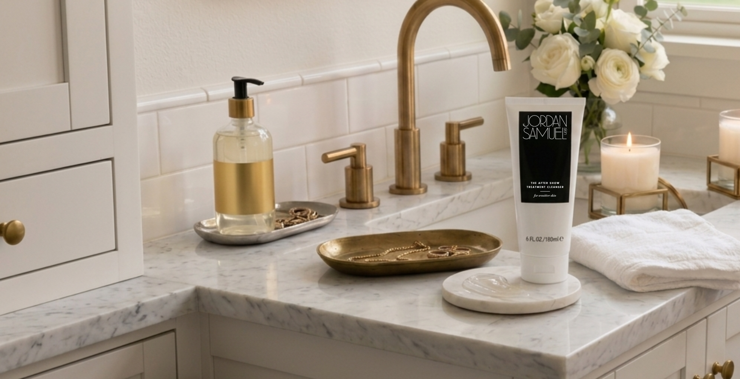 Jordan Samuel Skin's white tube of The After Show Treatment Cleanser for Sensitive Skin with black label stands on a white marble coaster on a white marble bathroom vanity with brass hardware. The cleanser tube is next a swatch of the cleanser showing its cushiony, jelly-like texture. A jewelry tray with gold necklace and rings, a lit white candle in a glass and brass holder and a bouquet of white roses in a clear glass vase decorate the bathroom vanity.
