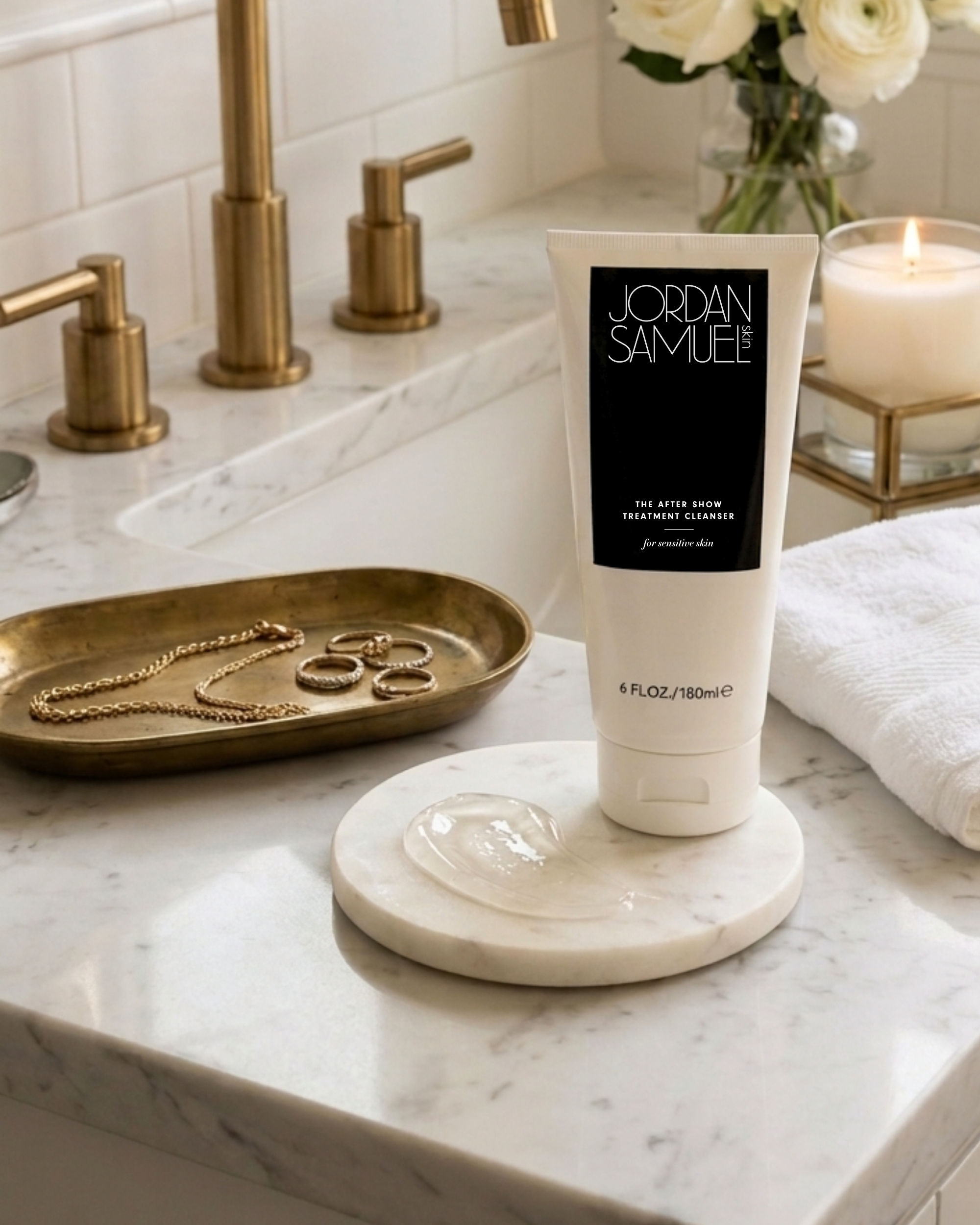 Jordan Samuel Skin's white tube of The After Show Treatment Cleanser for Sensitive Skin with black label stands on a white marble coaster on a white marble bathroom vanity with brass hardware. The cleanser tube is next a swatch of the cleanser showing its cushiony, jelly-like texture. A jewelry tray with gold necklace and rings, a lit white candle in a glass and brass holder and a bouquet of white roses in a clear glass vase decorate the bathroom vanity.