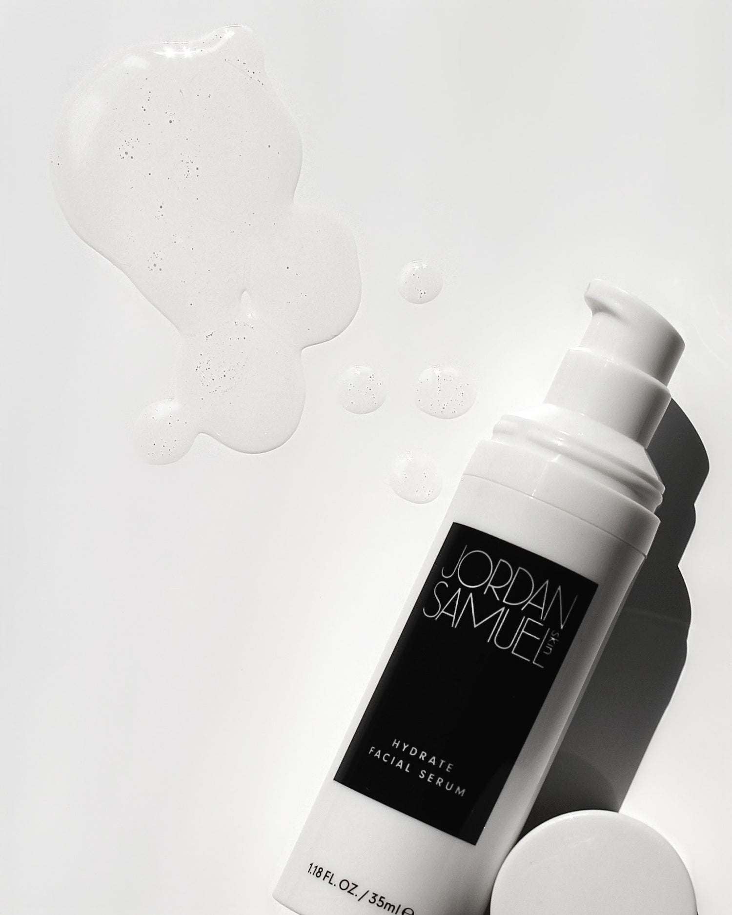 Hydrate Facial Serum's white airless pump bottle with black label without its cap on (the cap is resting next to the bottle) lays on a brightly lit white surface. The clear serum has been pumped on to the surface next to the bottle. 