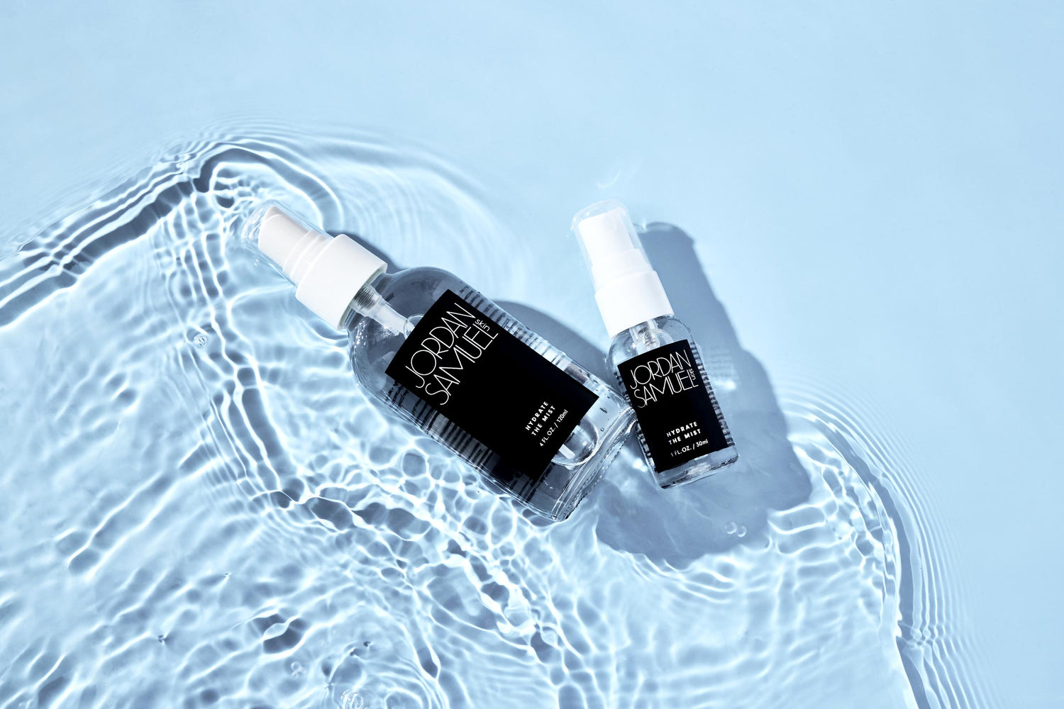 Hydrate the Mist and it's mini are laying in a shallow pool of light blue water that is seen swirling around the two bottles. 