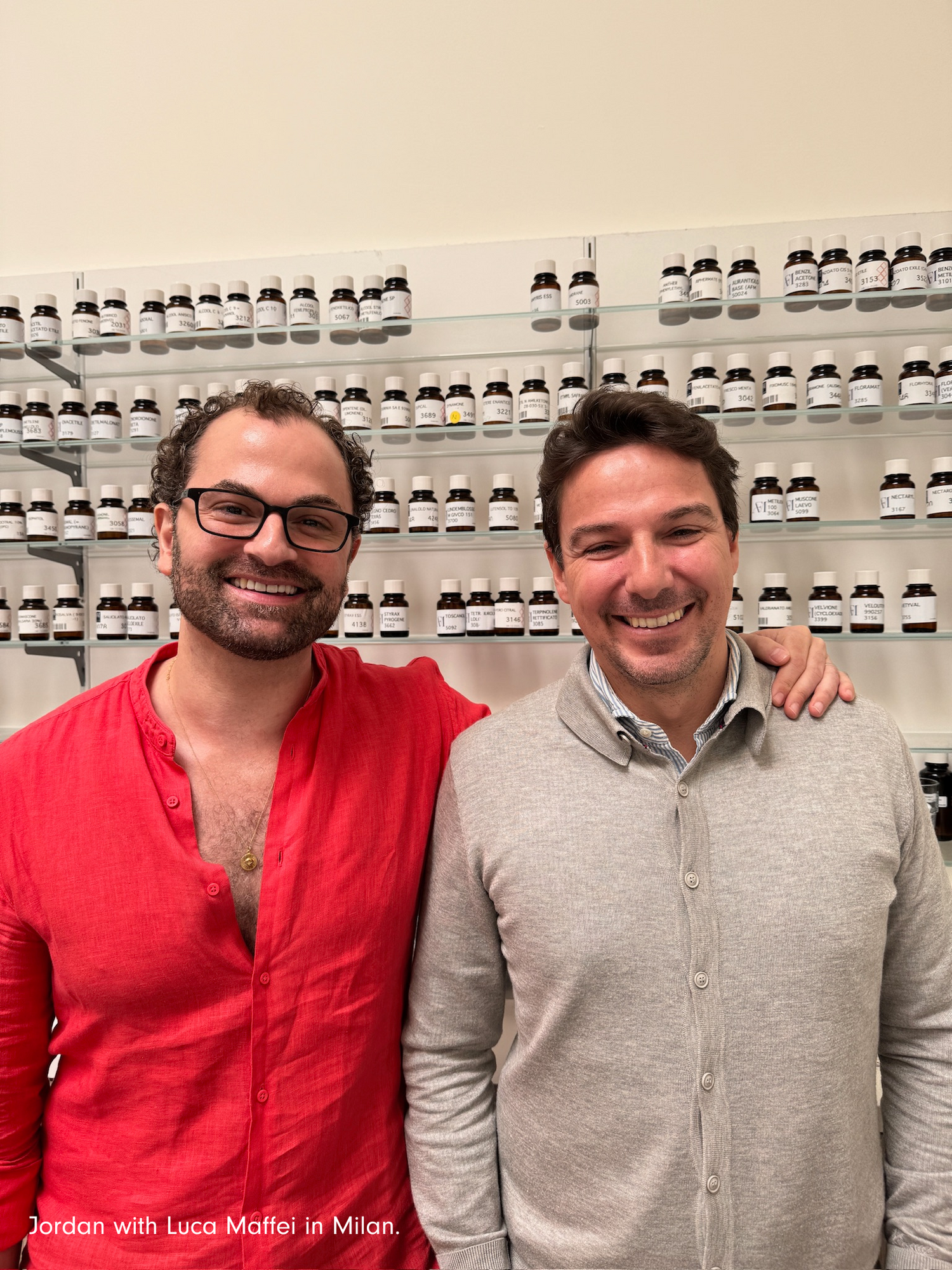Jordan wearing a red linen shirt next to Italian master perfurmer Lucca Maffei in a striped collared shirrt and khaki colored cardigan at Luccas Atelier in Milan. Bottles of fragrance notes line shelves in the background. 