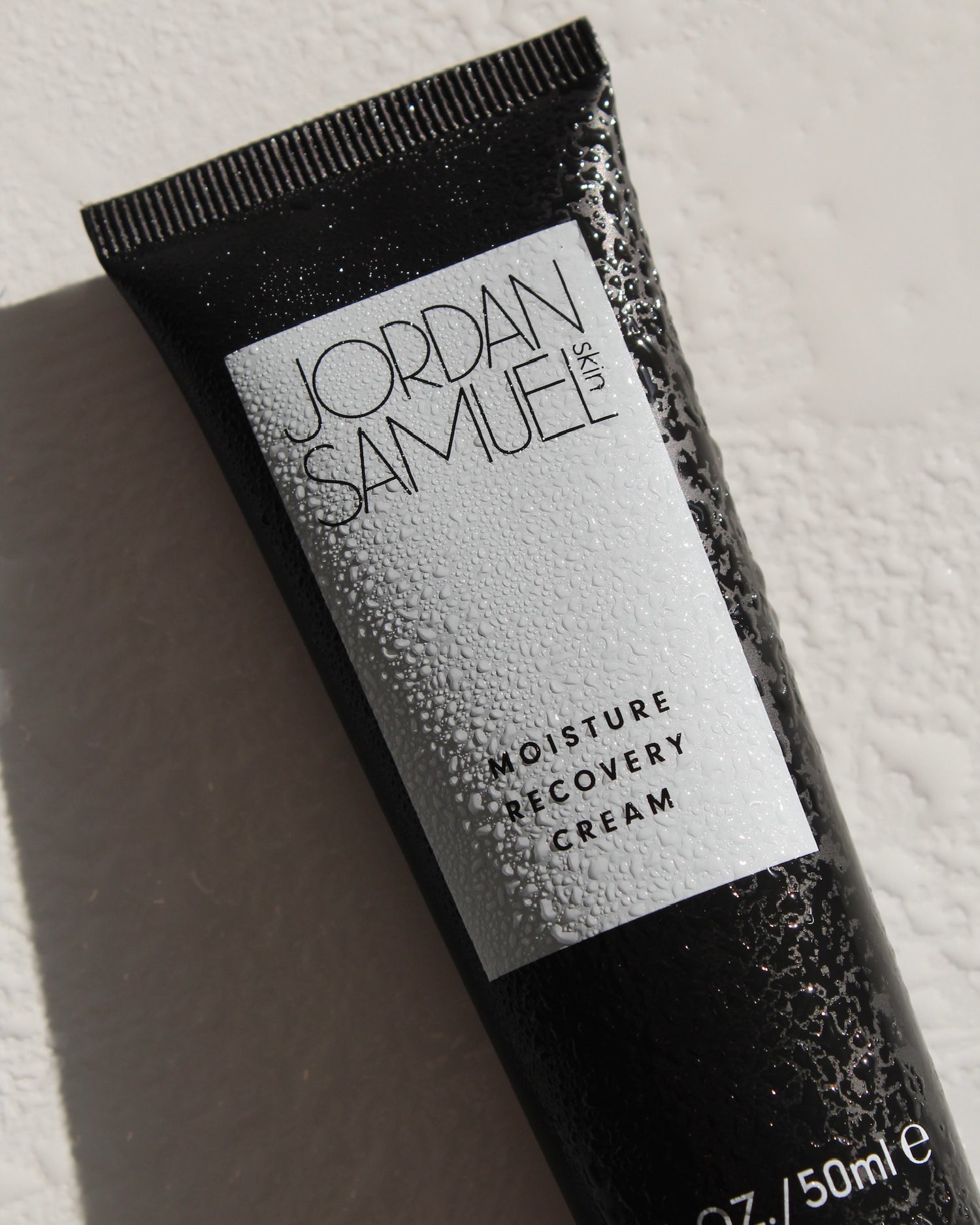 Aerial image of Moisture Recovery Cream's black tube with white label. Beads of water droplets cling to the tube. 