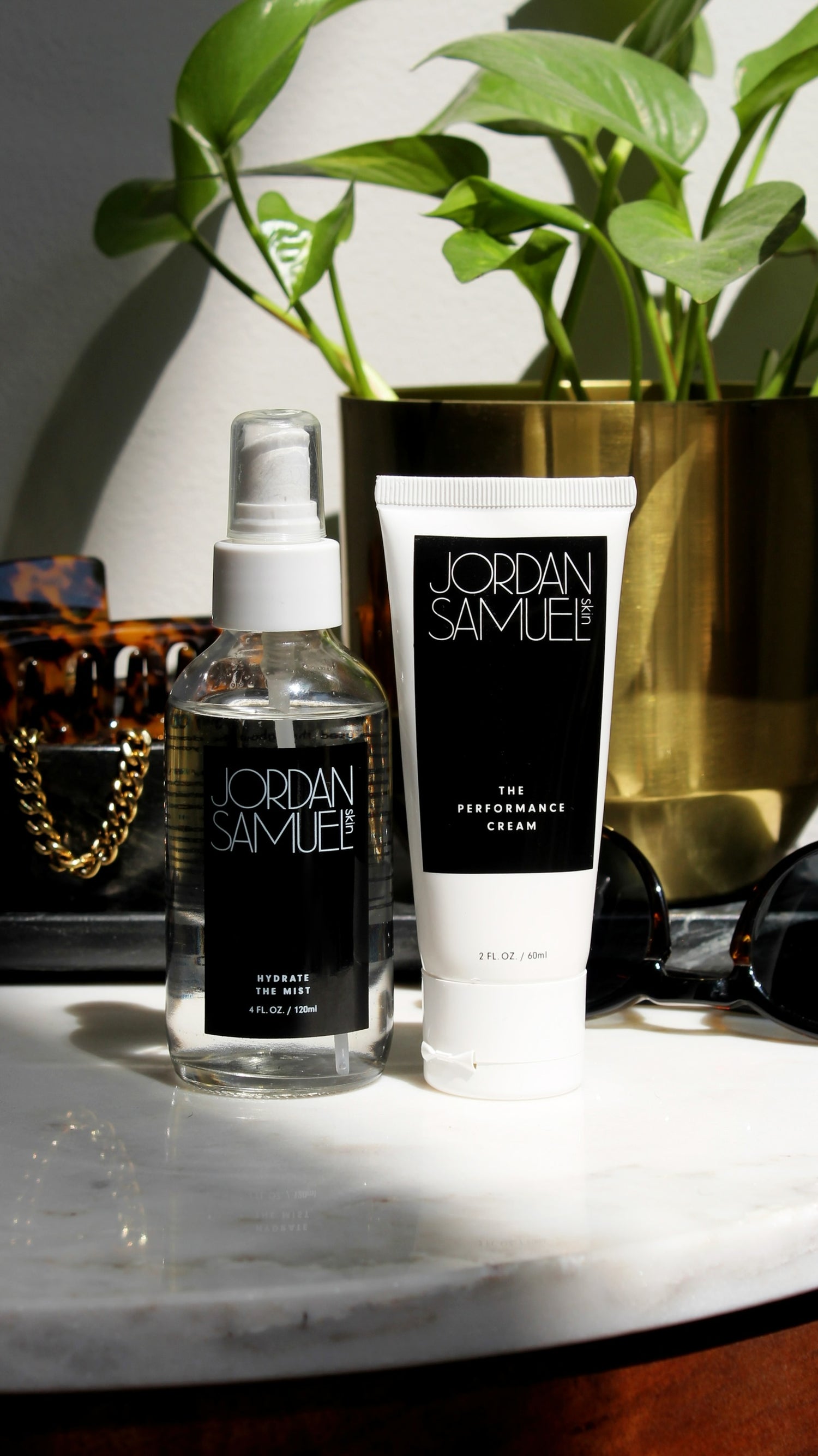The Performance Cream's white tube with black label and Hydrate the Mist's clear glass bottle with black label and white sprayer stand side by side on a white marble topped vanity with sunglasses, gold jewelry and a houseplant in gold pot. 