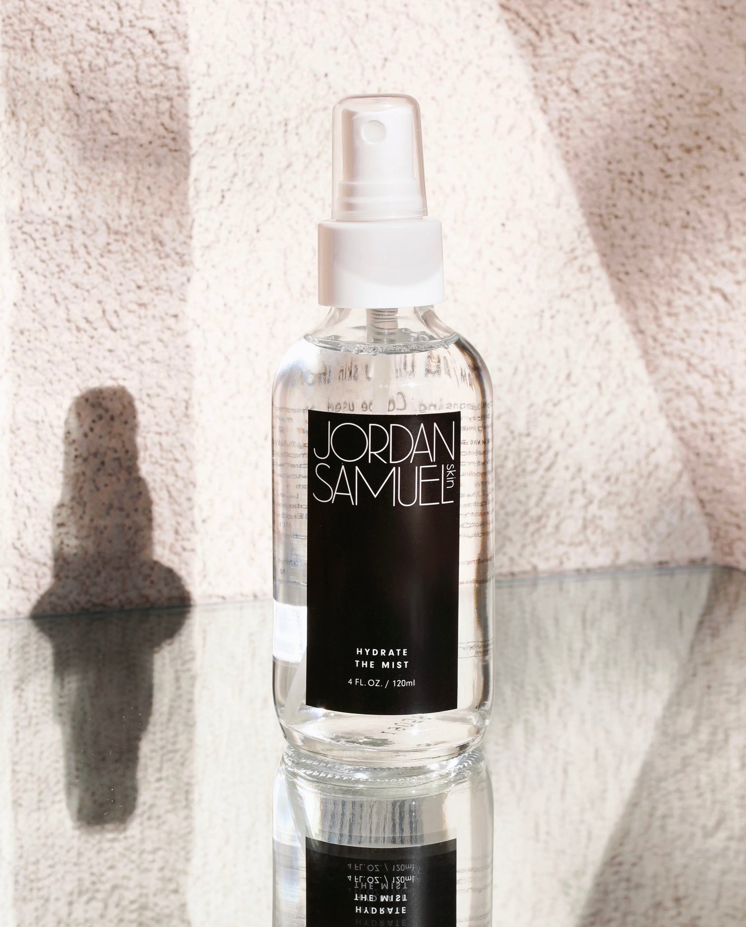 Hydrate the Mist's clear glass bottle with black label and white spray cap stand on a mirrored surface against a heavily textured white wall. 