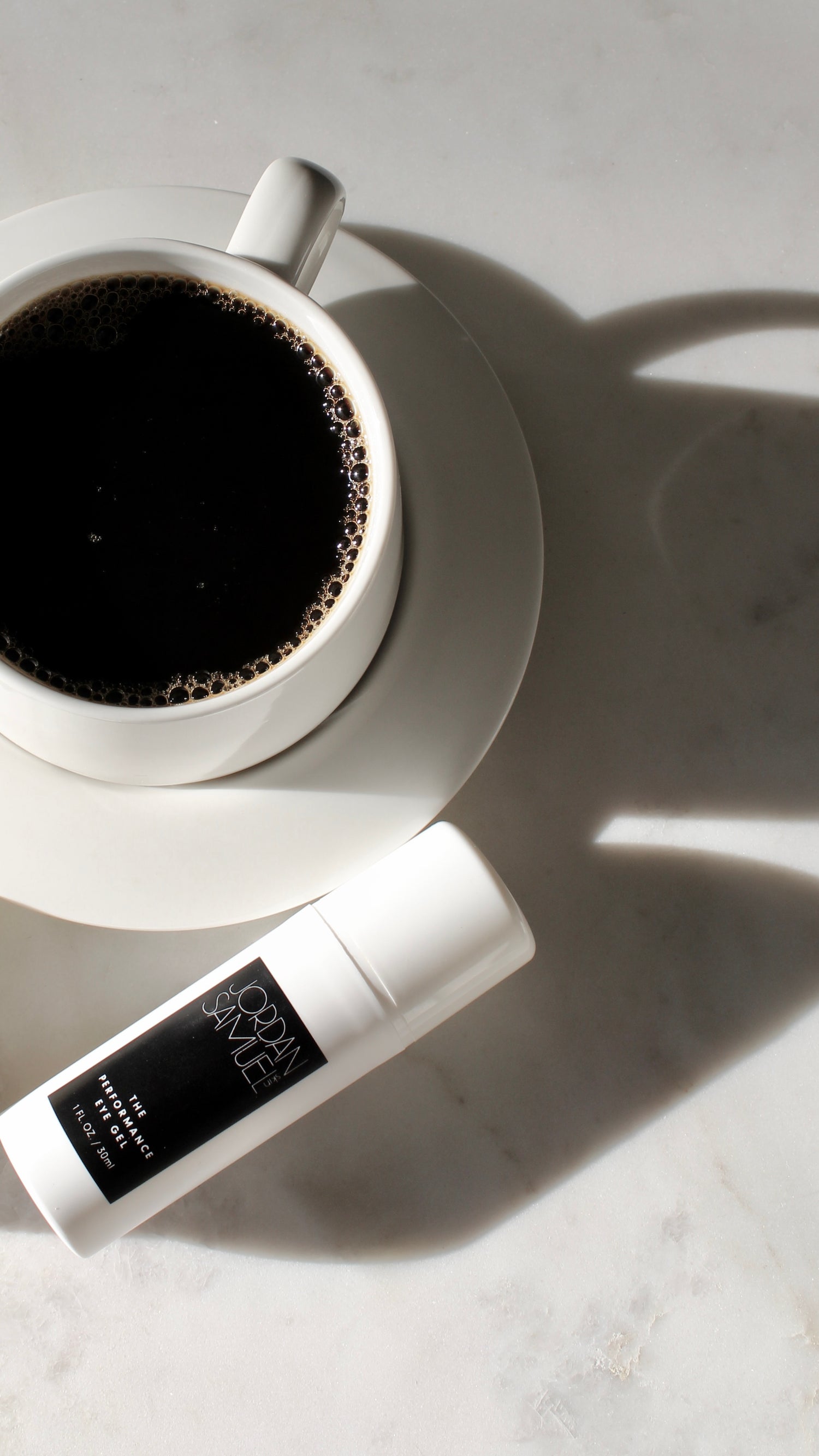The eye gel laying on a white marble table next to a white mug and saucer filled with black coffee.
