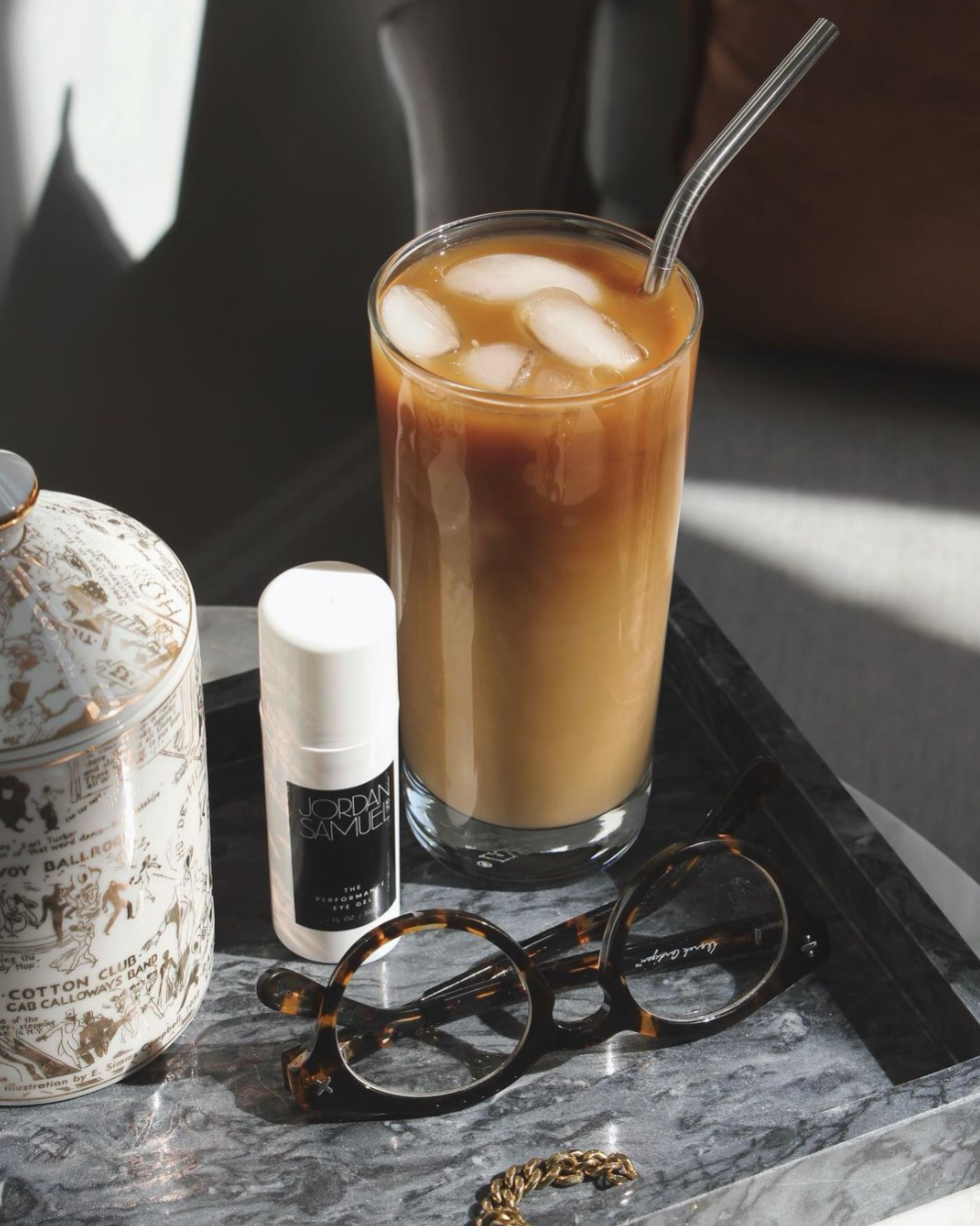 The Eye Gel's white airless pump bottle with black label stands on a grey stone tray next to a ceramic candle, gold jewelry, tortoise shell eyeglasses and a glass filled with iced coffee and a glass straw.