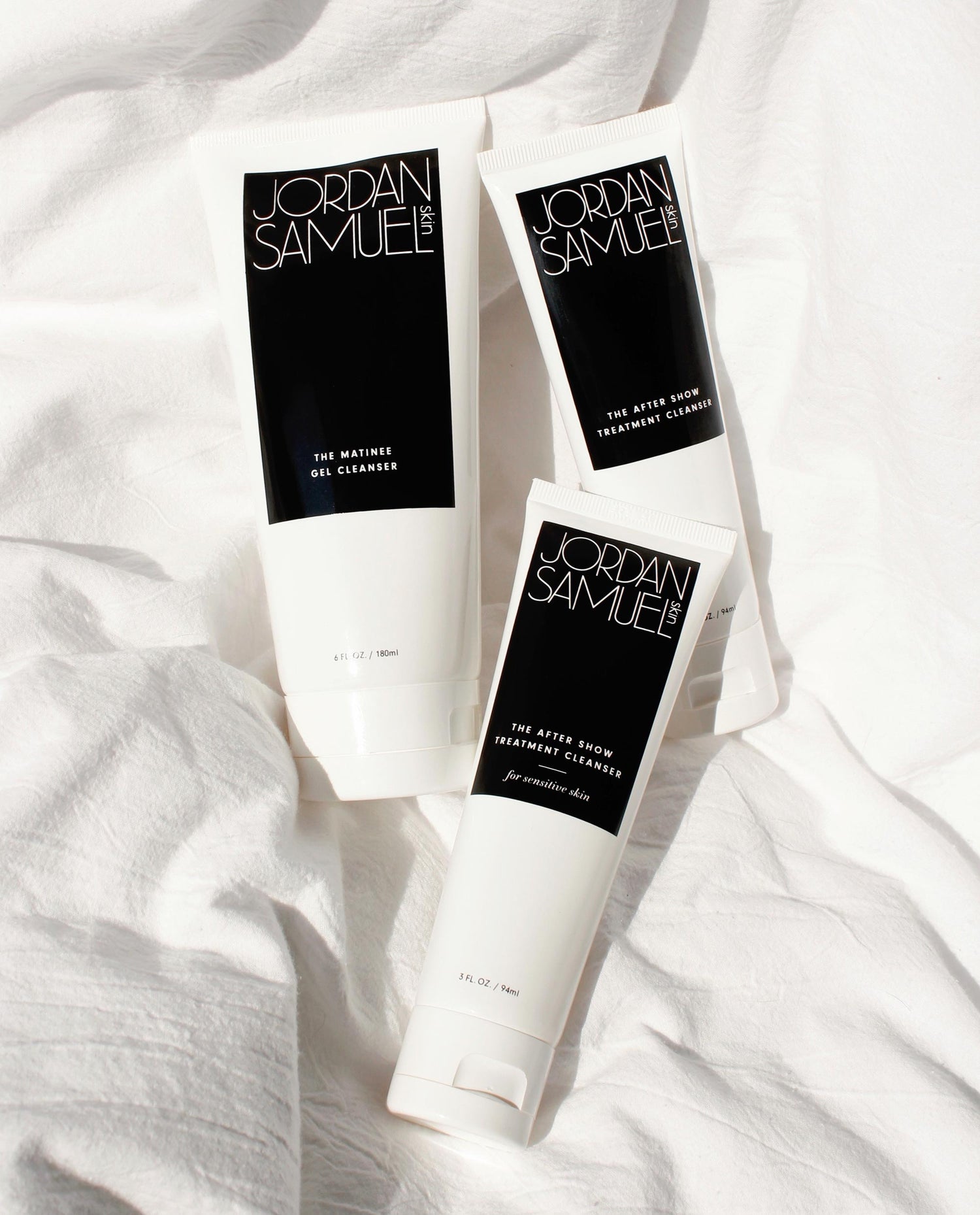3 JSS cleansers in white tubes with black labels grouped on a white linen duvet cover. 
